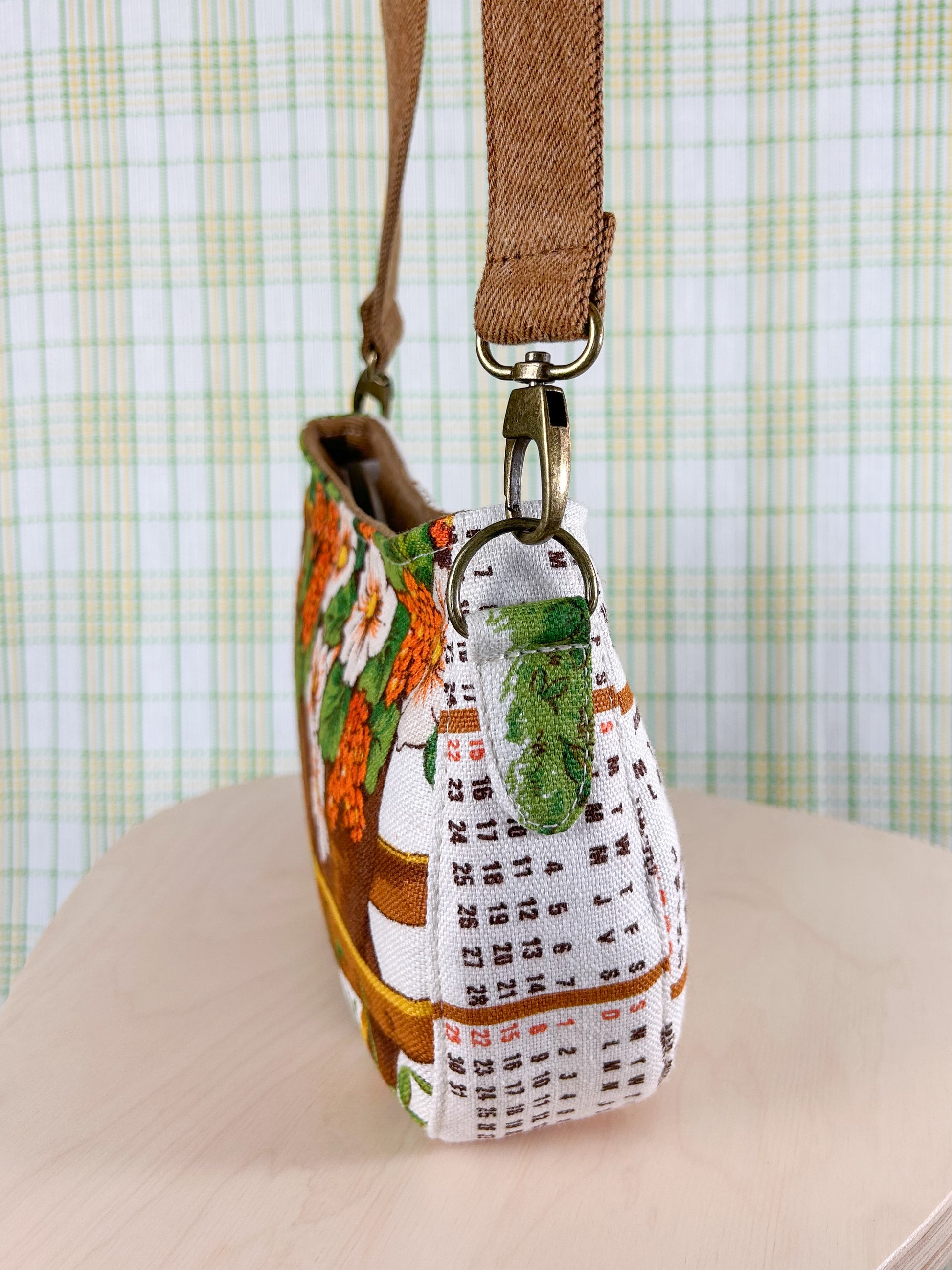 Side view of the handbag showing purse strap tabs, hardware and strap on a wooden stool in front of a plaid backdrop