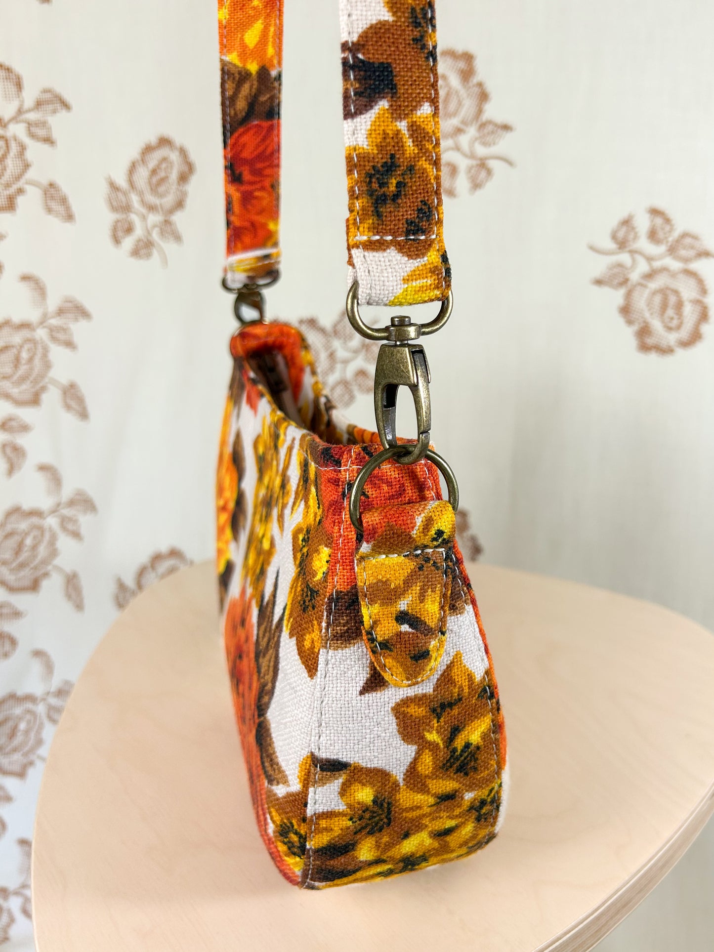 Side view of the handbag showing the purse strap tabs, antique brass hardware and strap on a wooden stool in front of a floral patterned backdrop