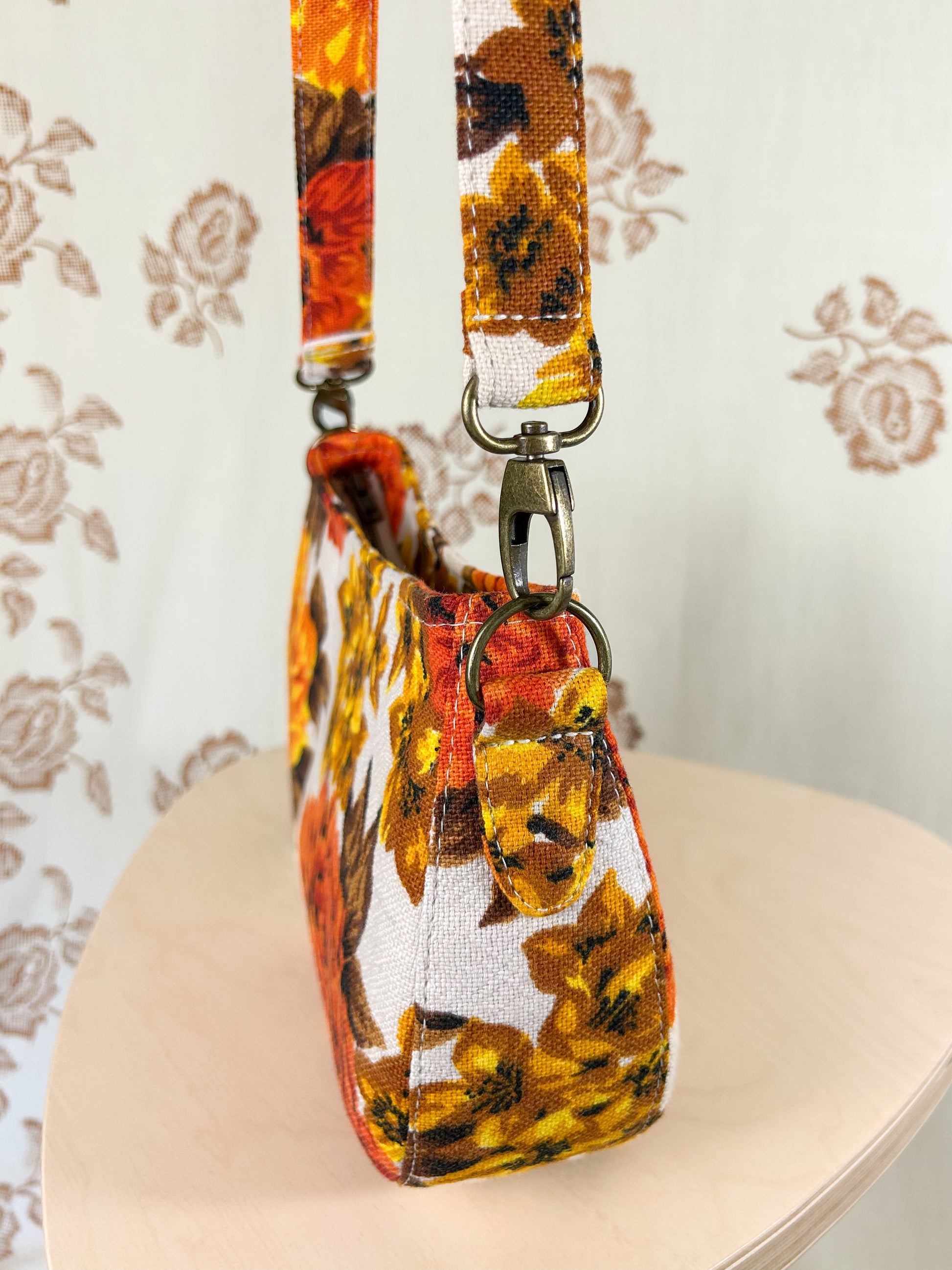 Side view of the handbag showing the purse strap tabs, antique brass hardware and strap on a wooden stool in front of a floral patterned backdrop