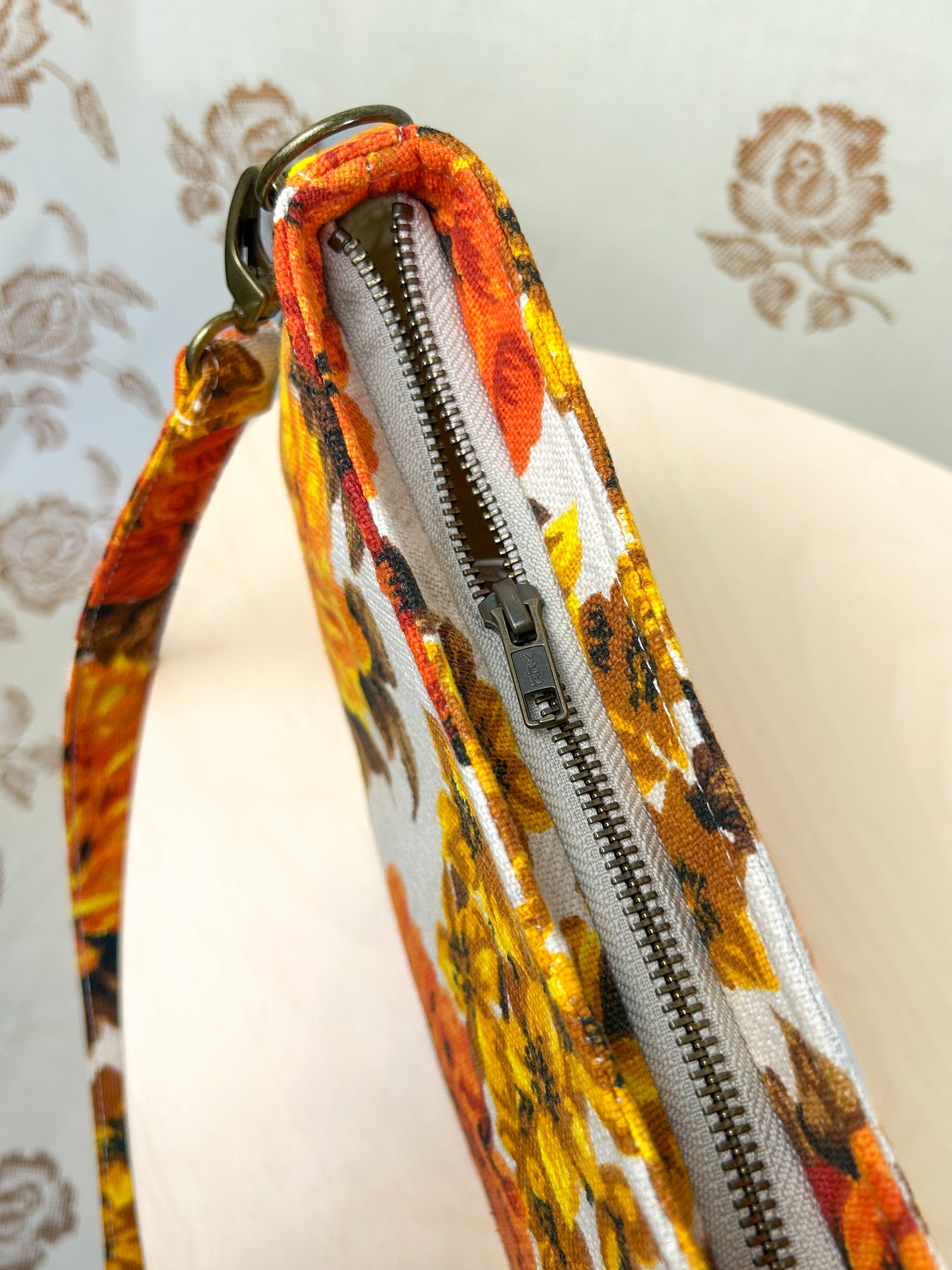 Top view of the handbag showing the antique brass zipper on a wooden stool in front of a floral patterned backdrop