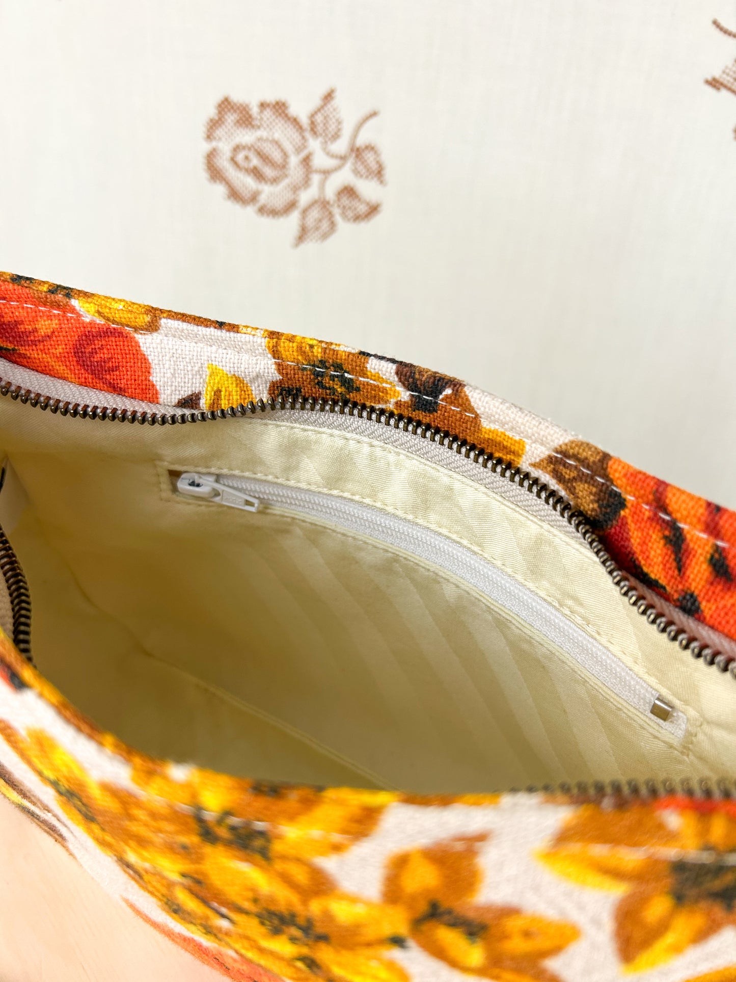 Interior view of the handbag showing the cream coloured lining and zippered pocket on a wooden stool in front of a floral patterned backdrop