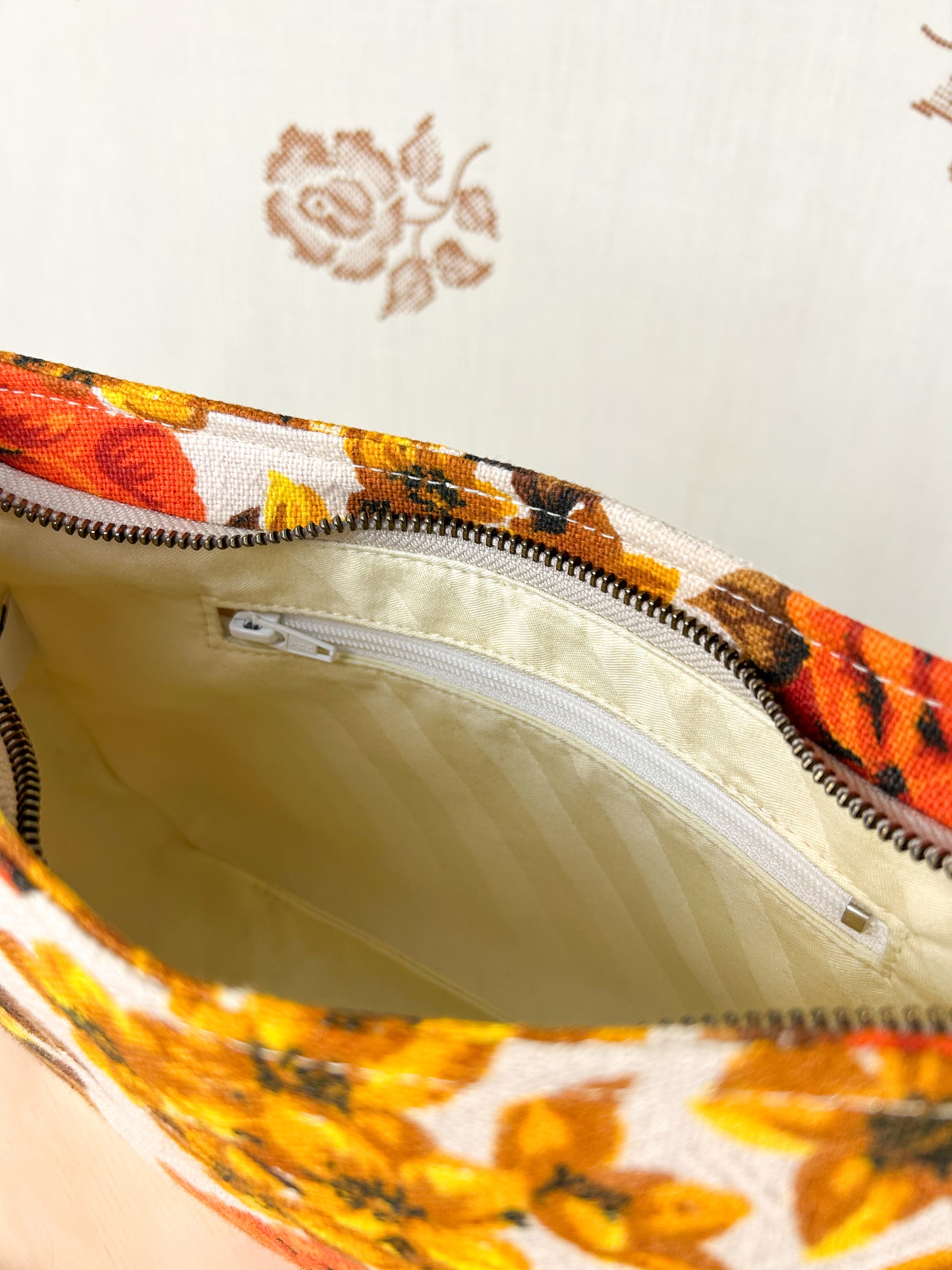 Interior view of the handbag showing the cream coloured lining and zippered pocket on a wooden stool in front of a floral patterned backdrop