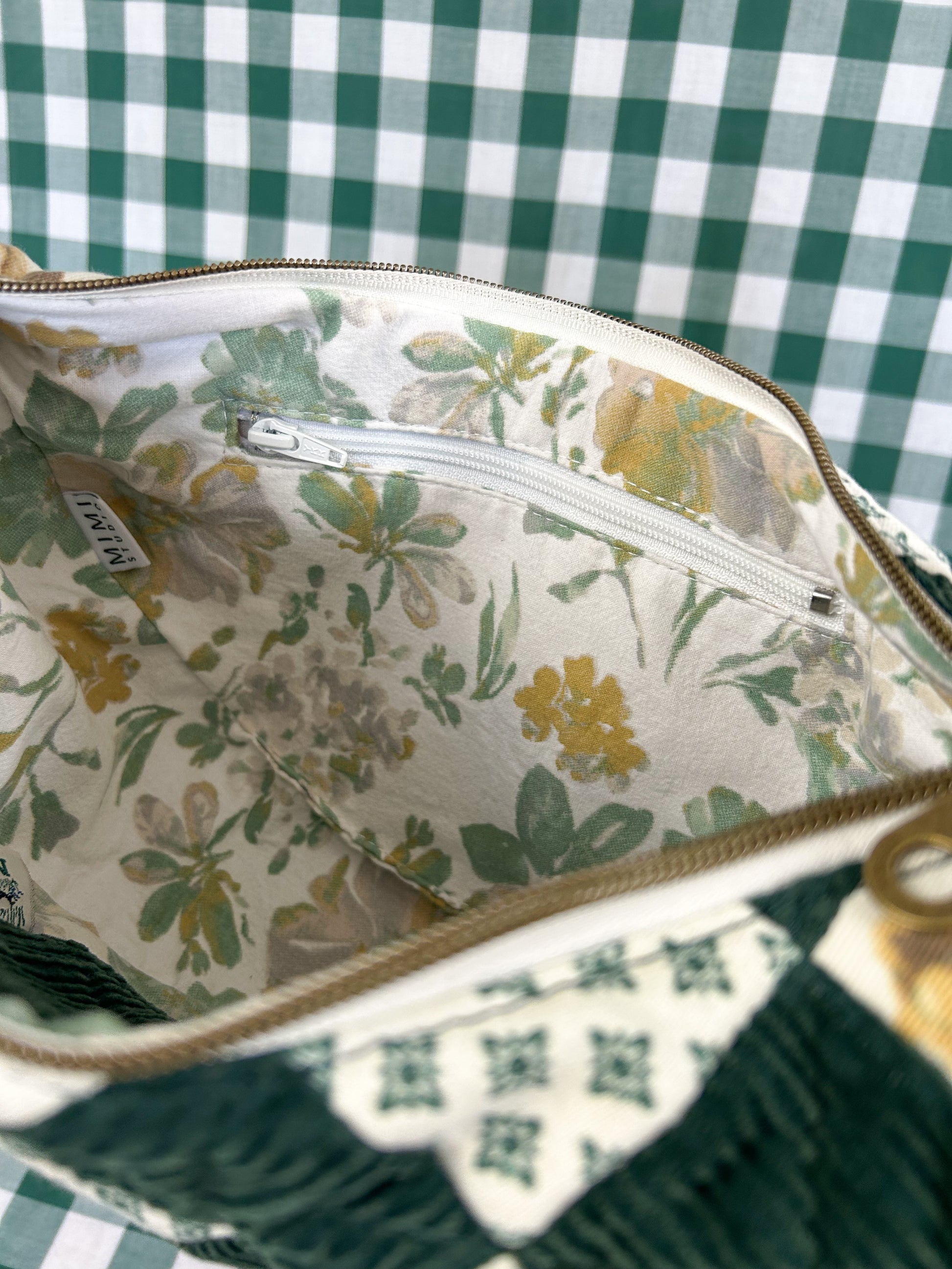 Interior view of the bag showing the green and white floral lining and zippered pocket in front of a green and white gingham backdrop
