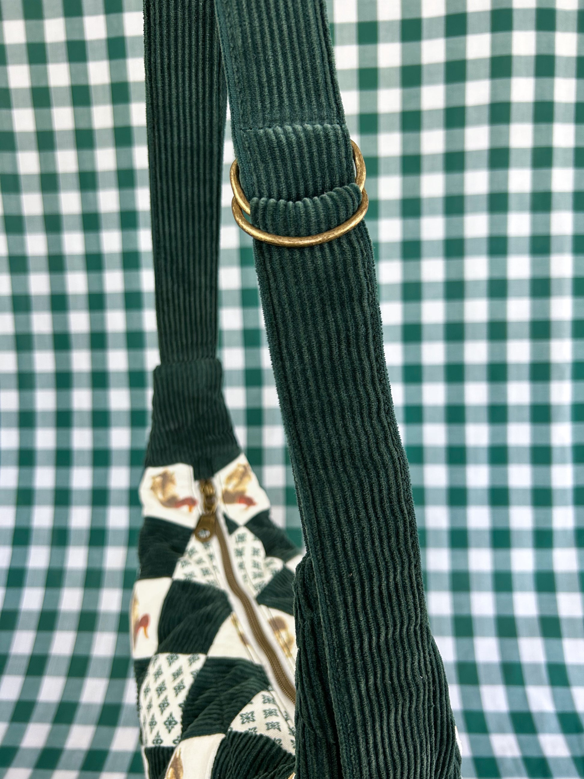 Green corduroy strap with d ring adjuster for the same crescent bag in front of a green and white gingham backdrop