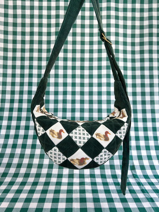 Dark green and duck patterned checkered quilted half moon crescent bag hanging in front of a green and white gingham backdrop