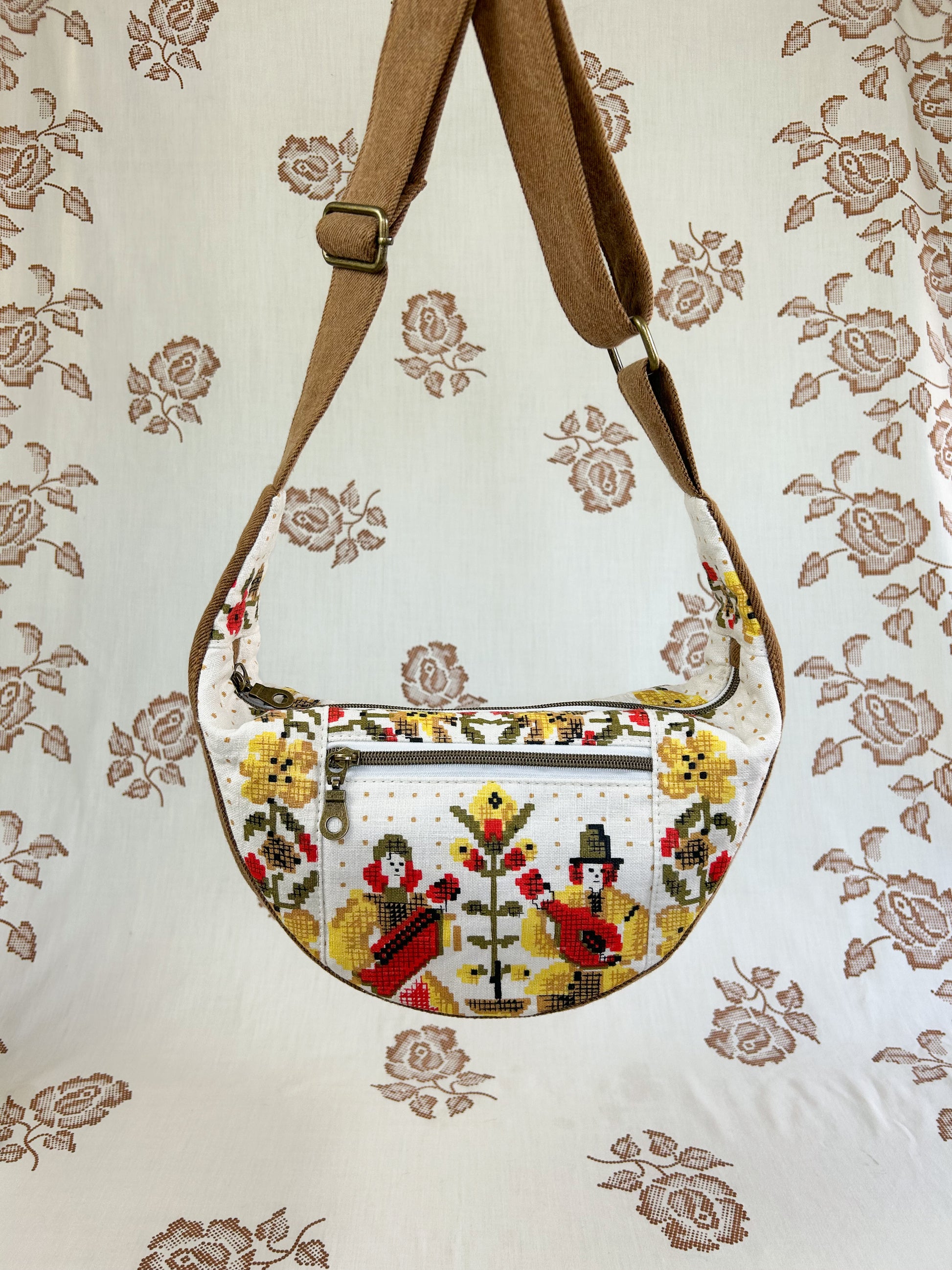 Half moon crescent bag with a yellow, red, green and ivory coloured musician and floral print hanging in front of a light cream coloured floral backdrop