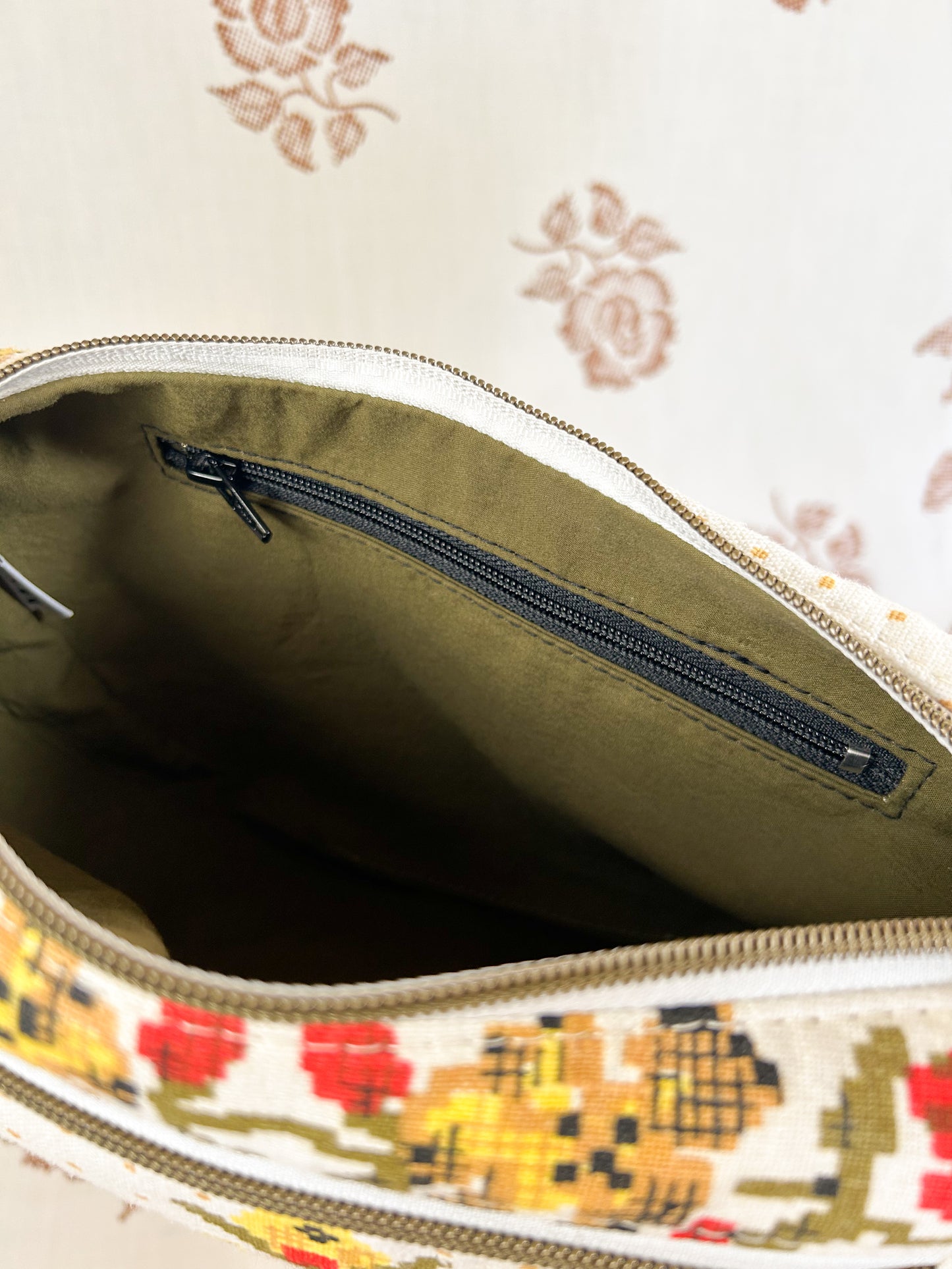 Interior view of the same bag showing the olive green lining and zippered pocket in front of a light cream coloured floral backdrop