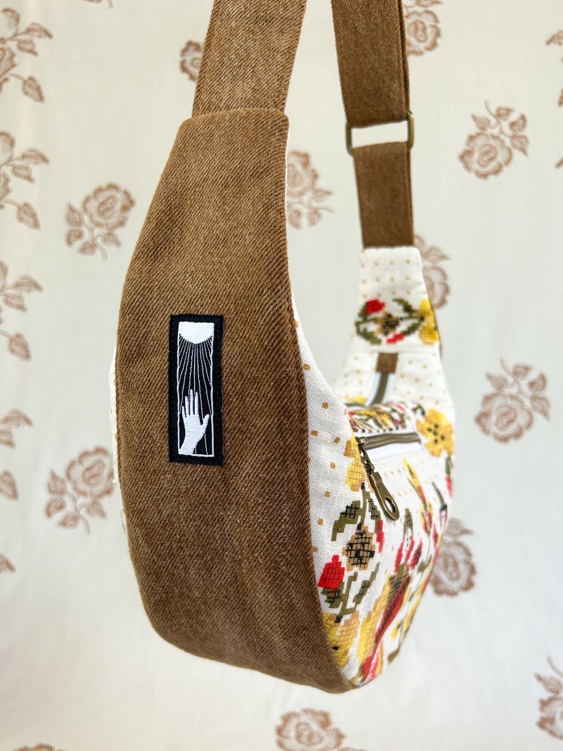 Side view of the same bag showing the tan coloured denim bottom and side piece and strap in front of a light cream coloured floral backdrop