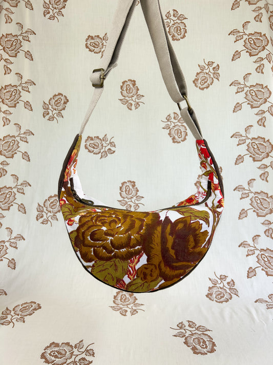 Brown, white, green, yellow and red floral printed half moon crescent bag hanging in front of a light cream coloured floral backdrop