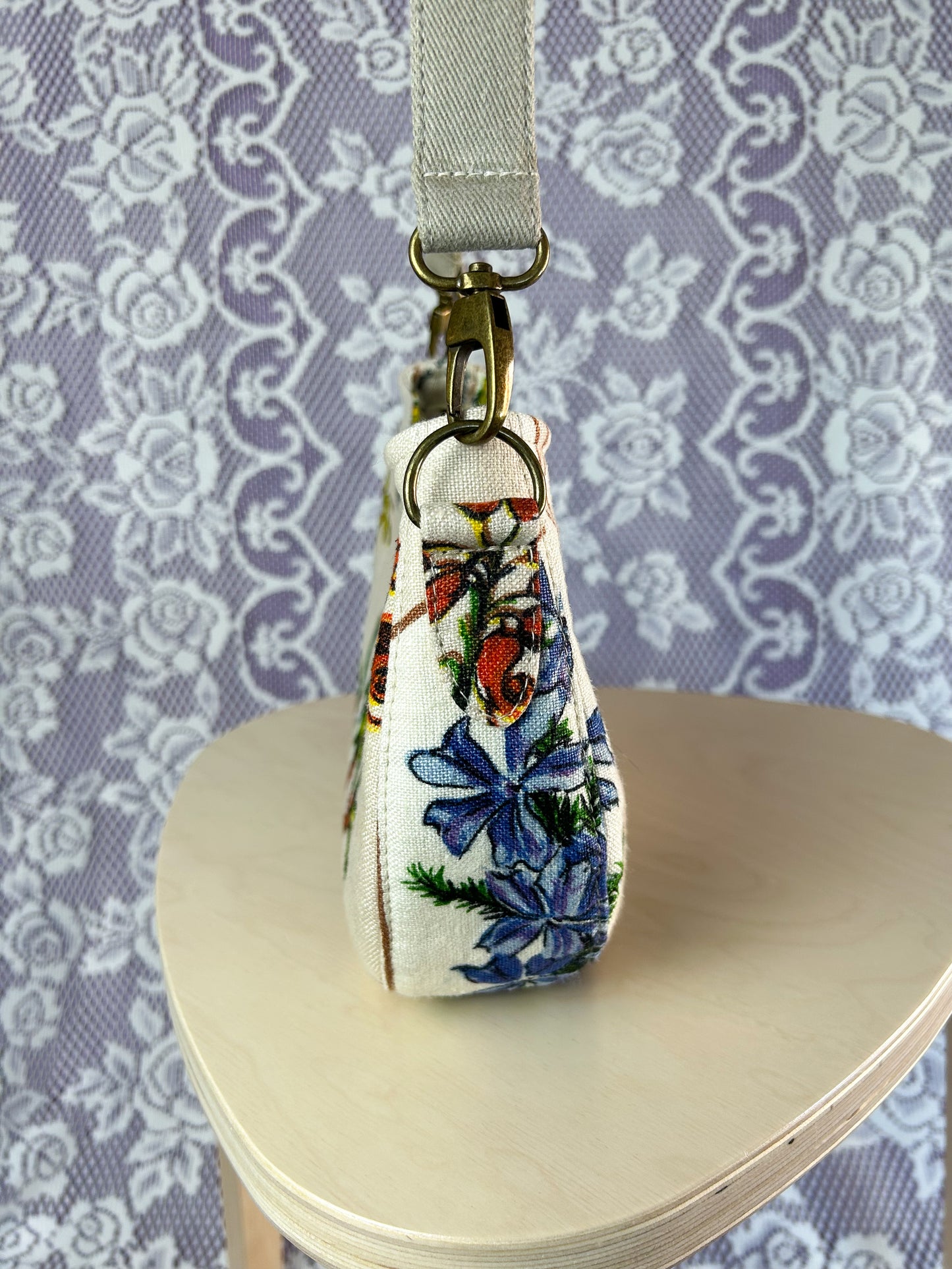 Side view of the handbag showing the purse strap tabs, hardware and strap on a wooden stool in front of a lace backdrop